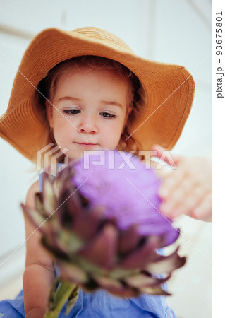 Cute little girl in straw hat touching big artichoke flower  93675801