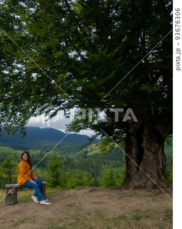 Female in casual clothes sits under large tree with forest background Female in casual clothes sits under large tree with forest background 93676306