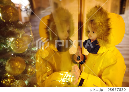 Little boy admires Christmas decorations in showcase of shop on winter evening. Tourist looking on Xmas tree toys and accessories on traditional Christmas market. Winter holidays in Europe. Little boy admires Christmas decorations in showcase of shop on winter evening. Tourist looking on Xmas tree toys and accessories on traditional Christmas market. Winter holidays in Europe. 93677820
