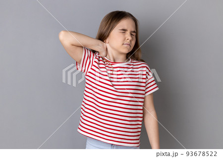 Portrait of unhealthy sick dark haired little girl wearing striped T-shirt standing massaging neck to relieve pain, muscle strain in back. Indoor studio shot isolated on gray background. 93678522