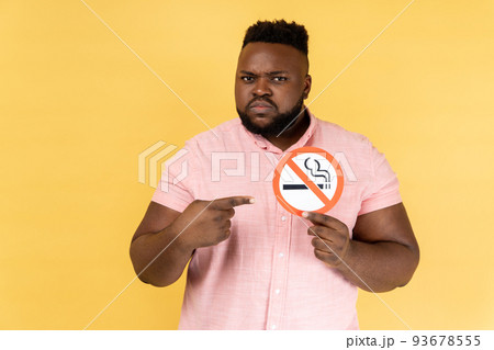 Portrait of serious man wearing pink shirt holding and pointing index finger at no smoking sign in hands, quit smoking, no tobacco day. Indoor studio shot isolated on yellow background. 93678555