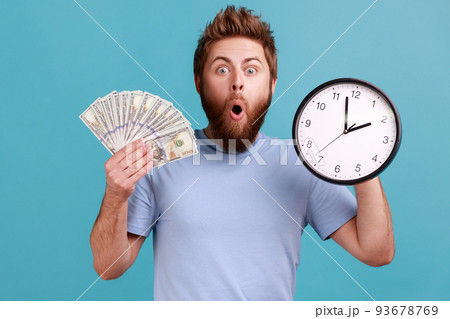 Portrait of surprised handsome bearded man holding big fan of dollar banknotes and wall clock, time is money, expressing astonishment. Indoor studio shot isolated on blue background. 93678769