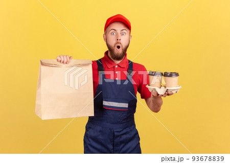 Portrait of astonished barded deliveryman wearing uniform holding paper package and take away coffee in hands, fast delivery, breakfast. Indoor studio shot isolated on yellow background. Portrait of astonished barded deliveryman wearing uniform holding paper package and take away coffee in hands, fast delivery, breakfast. Indoor studio shot isolated on yellow background. 93678839