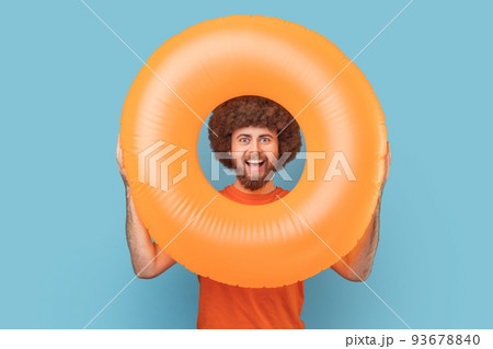 Portrait of man with Afro hairstyle with happy facial expression looking trough bright orange rubber ring, looking at camera, enjoying his vacation. Indoor studio shot isolated on blue background. Portrait of man with Afro hairstyle with happy facial expression looking trough bright orange rubber ring, looking at camera, enjoying his vacation. Indoor studio shot isolated on blue background. 93678840