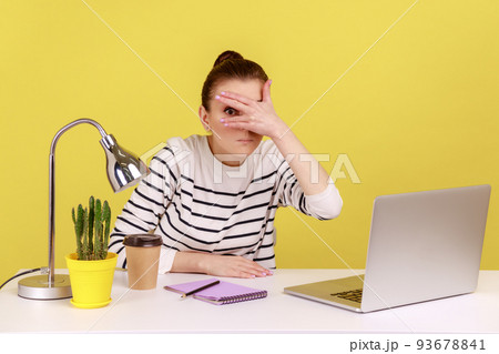 Curious woman manager sitting at workplace with laptop and spying, looking through fingers with interest, peeking secret gossip in office. Indoor studio studio shot isolated on yellow background. Curious woman manager sitting at workplace with laptop and spying, looking through fingers with interest, peeking secret gossip in office. Indoor studio studio shot isolated on yellow background. 93678841