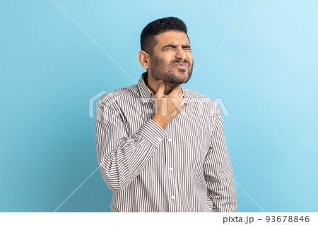 Throat pain. Portrait of unhappy ill businessman touching her neck, suffering sore throat, viral infection or flu symptoms, wearing striped shirt. Indoor studio shot isolated on blue background. Throat pain. Portrait of unhappy ill businessman touching her neck, suffering sore throat, viral infection or flu symptoms, wearing striped shirt. Indoor studio shot isolated on blue background. 93678846