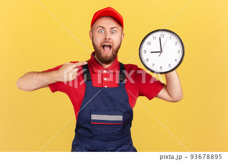 Shocked surprised man worker standing and pointing at big wall clock in his hands, looking at camera with open mouth, wearing overalls and red cap. Indoor studio shot isolated on yellow background. Shocked surprised man worker standing and pointing at big wall clock in his hands, looking at camera with open mouth, wearing overalls and red cap. Indoor studio shot isolated on yellow background. 93678895