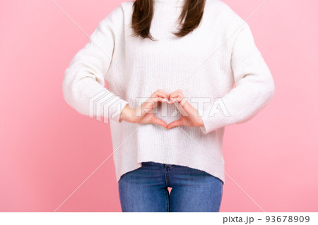Unknown girl making heart shape in front of her belly as symbol of women health care, healthy eating, wearing white casual style sweater and jeans. Indoor studio shot isolated on pink background. 93678909