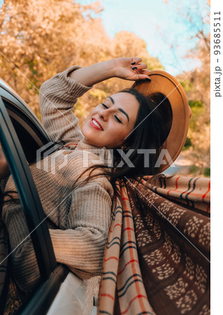 Beautiful young woman carrying hat and looking happy while enjoying road trip. Young woman with long hair in brown hat and sweater looking out from white car holding hat and scarf against autumn 93685511