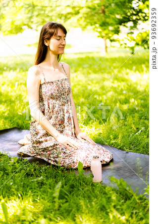 A beautiful young woman is sitting on a yoga mat in the park and doing yoga or meditation. Copy space. The concept fitness and a healthy lifestyle. 93692359