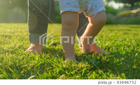 Walking children's bare feet on a green lawn close-up. Child learns to take the first steps on the grass. Baby learns to walk with the help of his mother on a green grass in the park. 93696752