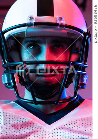 Close-up portrait of young man, american football player in helmet posing isolated over purple background neon light. Close-up portrait of young man, american football player in helmet posing isolated over purple background neon light. 93698426
