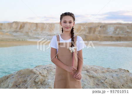 A beautiful teenage girl in a beige sundress is standing by the river in the mountains straightening her braids 93701051