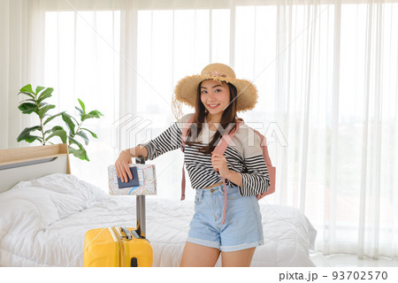 Young Asian woman in summer casual clothes, knapsack and straw hat standing with yellow suitcase in bedroom. Ready for holiday vacation trip. 93702570