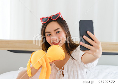 Young Asian woman show yellow.bikini while talking to facetime calling with her friend in bedroom. Using smartphone taking selfie on front camera . 93702572