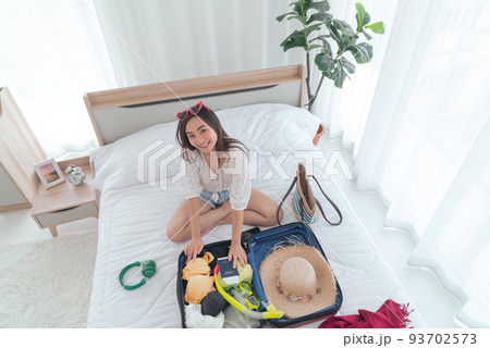 Young Asian woman looking at camera while packing summer clothes and beach accessories into suitcases on bed. Preparing travel items for new journey. summer holidays, vacation concept. 93702573