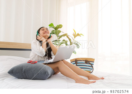 Young Asian woman wear green headphones relaxing and listening the music while using laptop computer on the bed, girl enjoys audio sound, relaxing 93702574