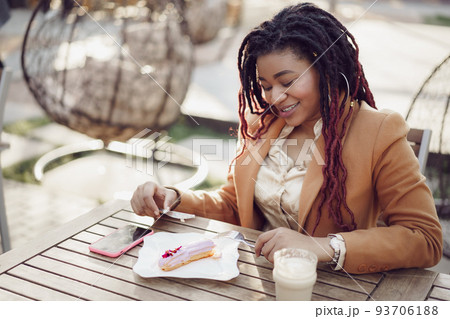 Smiling african american woman drinking coffee and eating dessert in outdoor cafe Smiling african american woman drinking coffee and eating dessert in outdoor cafe 93706188