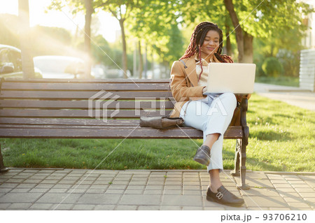 African american woman freelancer sitting on a bench in a street and using laptop African american woman freelancer sitting on a bench in a street and using laptop 93706210
