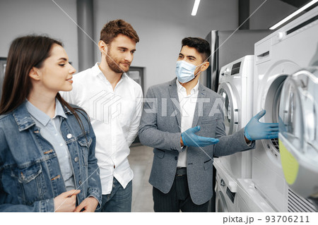 Salesman in hypermarket wearing medical mask demonstrates his clients a new washing machine 93706211