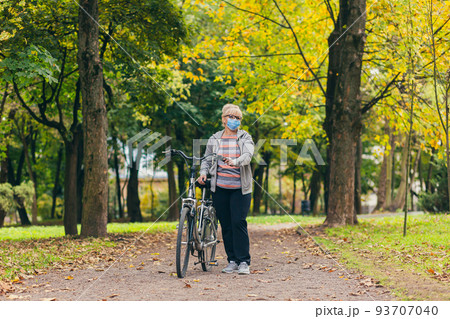 Senior beautiful blonde woman rides a bike in the park, in the woods, in nature in a medical mask during quarantine, leads a healthy active lifestyle 93707040