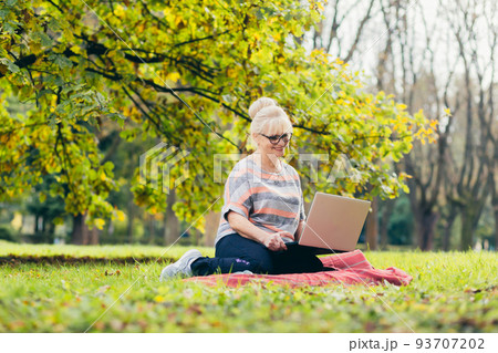 Beautiful blonde woman, senior, sitting on the grass in the park uses a laptop, works, studies, talks on video link Beautiful blonde woman, senior, sitting on the grass in the park uses a laptop, works, studies, talks on video link 93707202