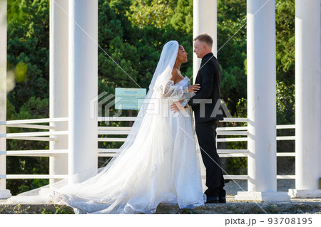 Mixed-racial newlyweds on a walk hugging against the backdrop of a beautiful gazebo 93708195
