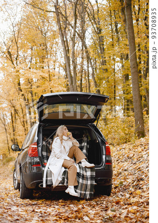 Blonde woman sitting in a black car trunk in autumn forest. Woman wearing white coat. Girl sitting on a plaid blanket. 93708935