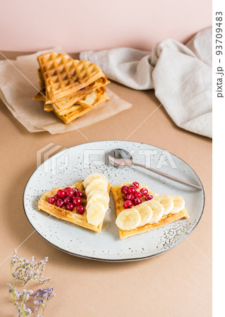 Heart shaped Belgian waffles with red berry and banana on a plate and a stack of waffles on the table. Valentine's Day. Vertical view Heart shaped Belgian waffles with red berry and banana on a plate and a stack of waffles on the table. Valentine's Day. Vertical view 93709483