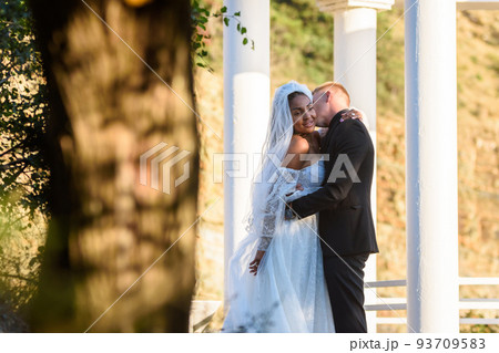 Young beautiful interracial newlyweds hugging against the background of white columns Young beautiful interracial newlyweds hugging against the background of white columns 93709583