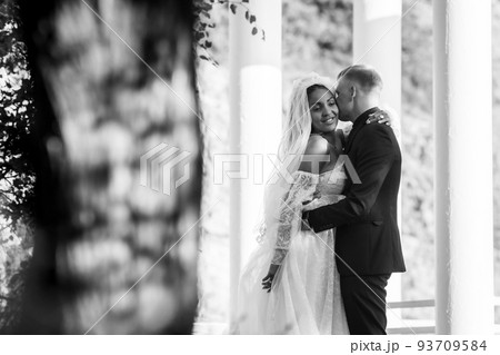 Portrait of interracial newlyweds hugging against the background of columns, black and white photography Portrait of interracial newlyweds hugging against the background of columns, black and white photography 93709584