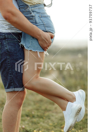 The guy holds the girl with strong hands against the background of a beautifully blurred field. She has long slender legs in white sneakers. 93709737