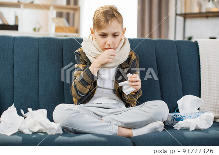 Pleasant tired sick teen boy with scarf around neck, sitting on soft couch and coughing Pleasant tired sick teen boy with scarf around neck, sitting on soft couch and coughing 93722326