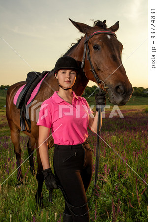 Beautiful smiling girl jockey stand next to her brown horse wearing special uniform on a sky and green field background on a sunset. 93724831