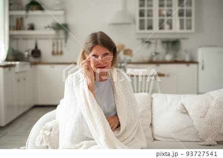 An elderly woman in glasses in a plaid is warming herself on the sofa at home. 93727541