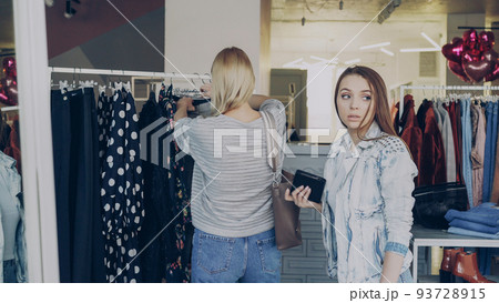 Young woman stealing wallet from female customer choosing clothes in shop. Thief looking around carefully and walking away. Fine boutique with clothing, bags and shoes in background 93728915