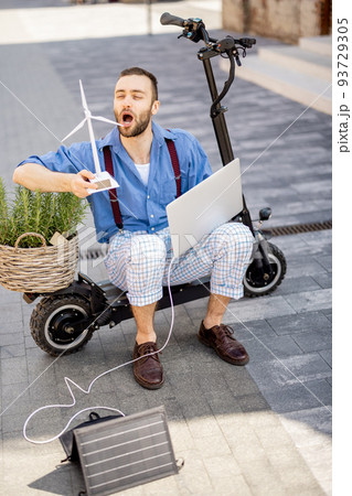 Man blows on toy wind generator while sitting with laptop and green plant on electric scooter outdoors Man blows on toy wind generator while sitting with laptop and green plant on electric scooter outdoors 93729305