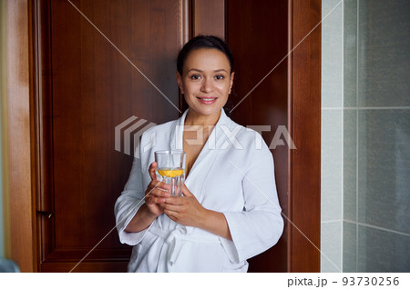 Close-up portrait. Beautiful attractive Hispanic woman with glowing skin, in bathrobe, smiles looking at camera, holds glass with lemon water for good start to the day. Diet, vitality, healthy habits Close-up portrait. Beautiful attractive Hispanic woman with glowing skin, in bathrobe, smiles looking at camera, holds glass with lemon water for good start to the day. Diet, vitality, healthy habits 93730256