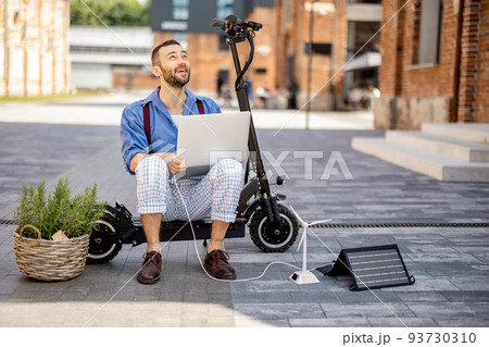 Man charges laptop with solar panels while sitting on electric scooter outdoors Man charges laptop with solar panels while sitting on electric scooter outdoors 93730310