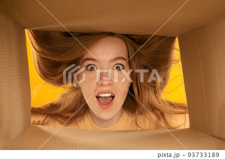 Closeup young girl's face. Young exressive girl looking inside carton box during opening, unpacking it. Emotions, black friday, holidays, online shopping concept Closeup young girl's face. Young exressive girl looking inside carton box during opening, unpacking it. Emotions, black friday, holidays, online shopping concept 93733189