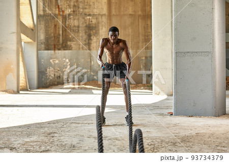 Rope workout. Sport man doing battle ropes exercise outdoor. Black male athlete exercising, doing functional fitness training with heavy rope 93734379