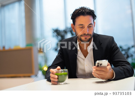Portrait of happy handsome Hispanic businessman at coffee shop having green tea while using phone Portrait of happy handsome Hispanic businessman at coffee shop having green tea while using phone 93735103