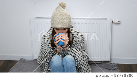 Frozen woman drinking tea at home. Young woman in hat, an warm blanket, near radiator try warm herself with drink in white room. Cold house, poor central heating, low temperature in apartment concept  93738880