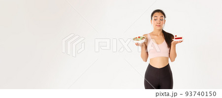 Active lifestyle, fitness and wellbeing concept. Indecisive cute asian slim girl in workout outfit, holding healthy salad and cake, hesitating, need stay on diet, losing weight, white background 93740150