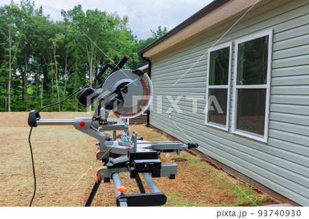 With a circular miter saw on a saw stand, a newly constructed house can be seen under construction With a circular miter saw on a saw stand, a newly constructed house can be seen under construction 93740930