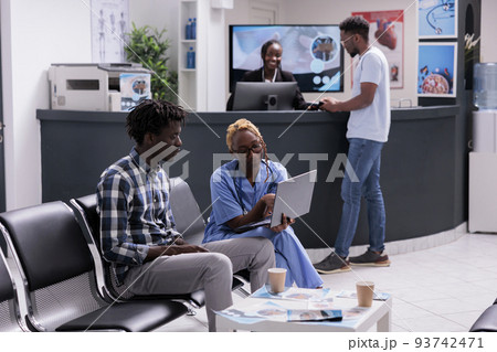 Medical assistant consulting patient at clinic, sitting in wating room lobby and using laptop. Explaining diagnosis to give recovery treatment with healthcare insurance and support, waiting area. 93742471