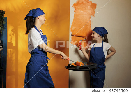 Mother and child painting room walls with orange color paint to renovate apartment interior with tools. Small family redecorating house room with paintbrush roller, home renovation. Mother and child painting room walls with orange color paint to renovate apartment interior with tools. Small family redecorating house room with paintbrush roller, home renovation. 93742500