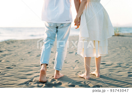 Man and woman stand on the sand barefoot holding hands. Cropped 93743047