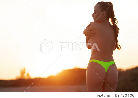 Sensual woman in bikini standing on sandy beach and covering breast at sunset 93750406