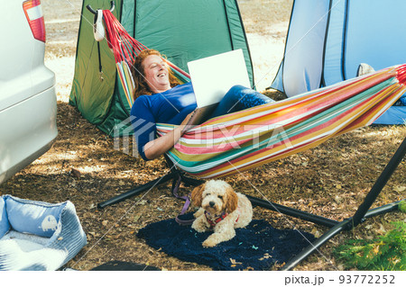 Adult woman working on laptop pc while lying in hammock, with her cockapoo puppy near motorhome on camping trip. Female living on camper car with animals and travel the world. Remote worker people 93772252